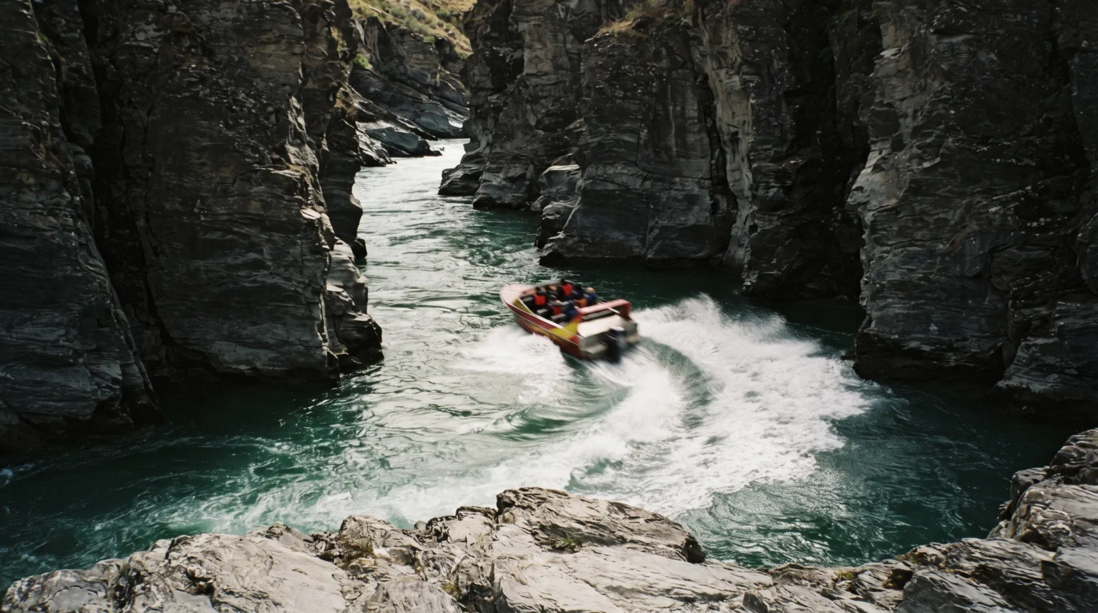 A red jet boat throwing spray as it turns hard between the grey schist walls of the Shotover Canyon on the Shotover Jet, Queenstown, New Zealand